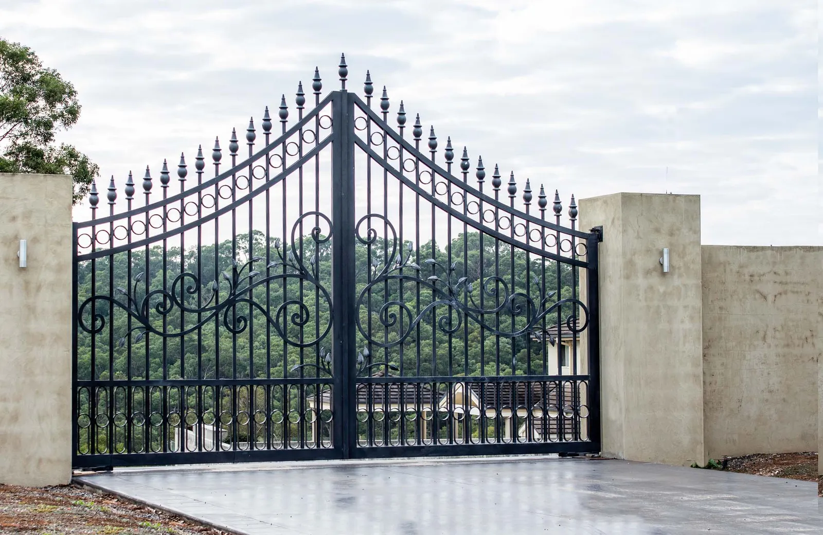 Black metal wrought iron driveway property entrance gates set in concrete brick fence, lights, garden trees in background